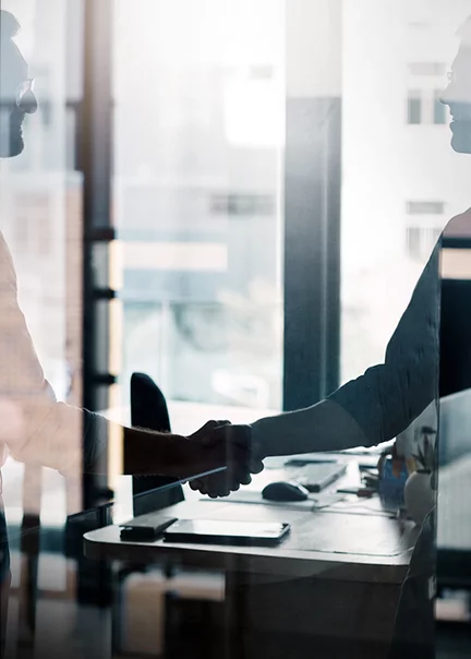 Shot through the reflections of a glass wall into an office where you can grasp the silhouttes of two men shaking hands