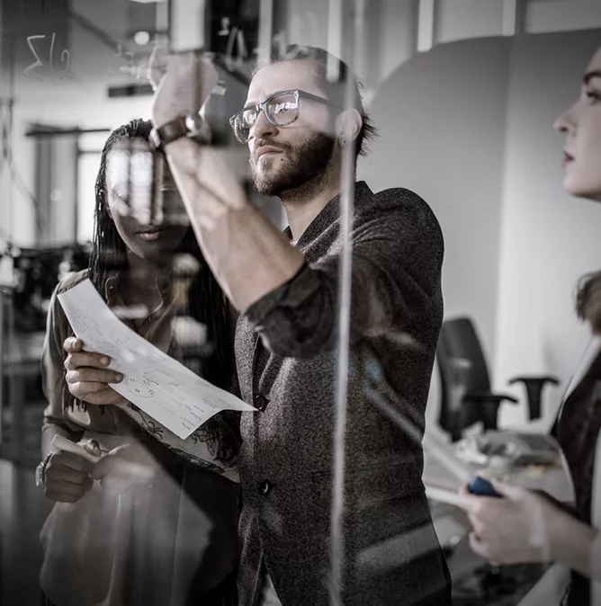 a man and two woman in a work meeting; the man writes formulas on a glass board, while the women examine the result